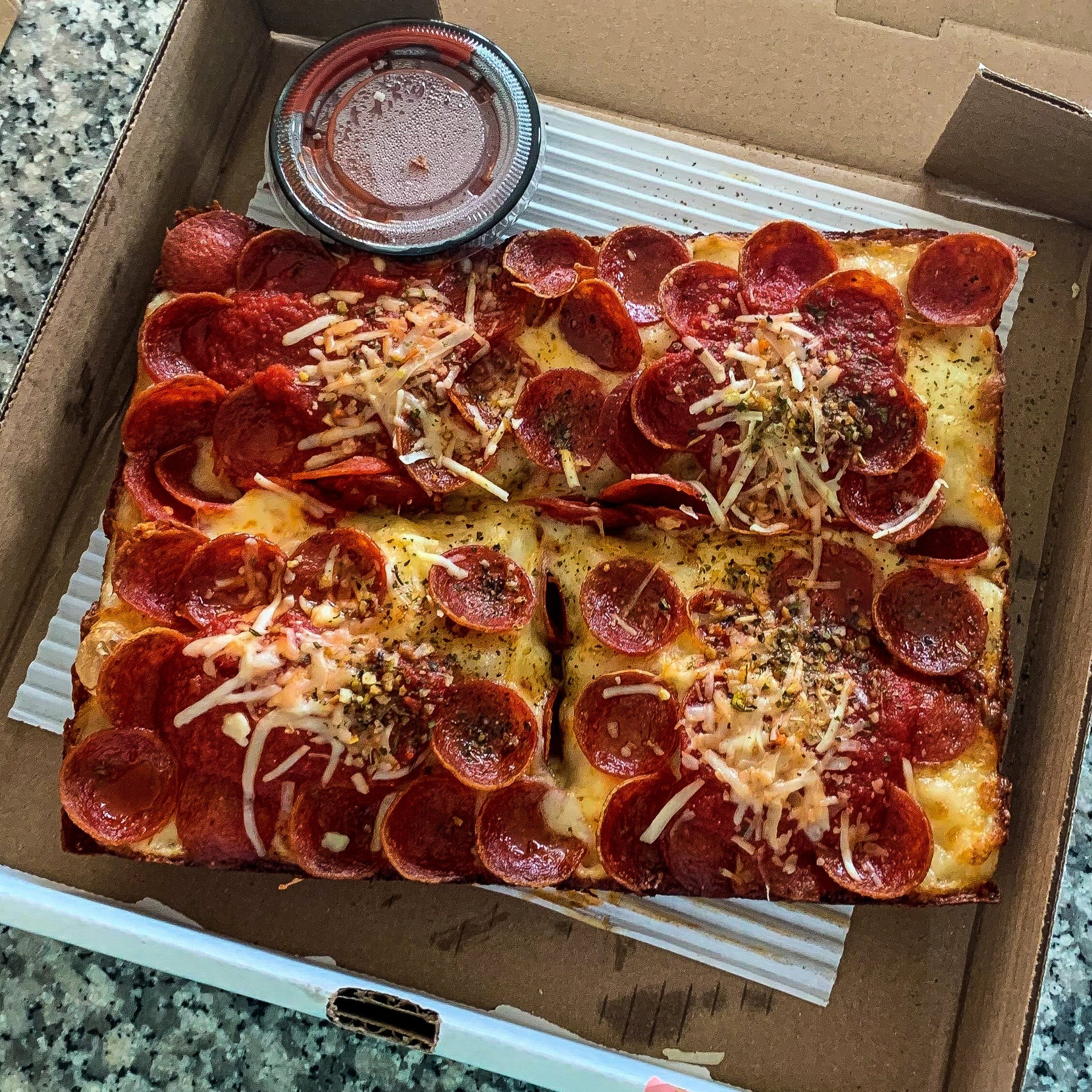 Close-up of a Detroit-style pepperoni pizza with crispy, caramelized cheese edges, served in a takeout box with a side of marinara sauce.