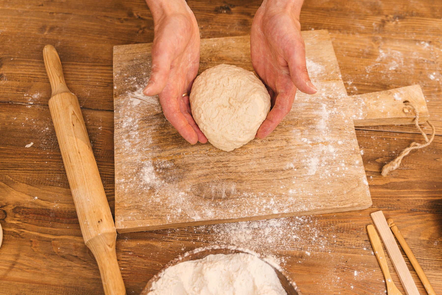 Hands shaping fresh pizza dough on a rustic wooden board, surrounded by flour and baking tools.