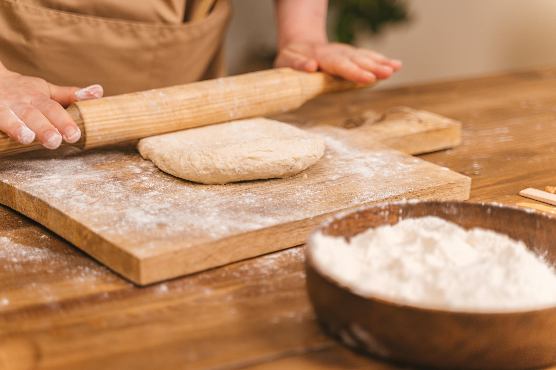 Close-up of hands using a rolling pin to flatten pizza dough on a floured wooden board, preparing it for baking.