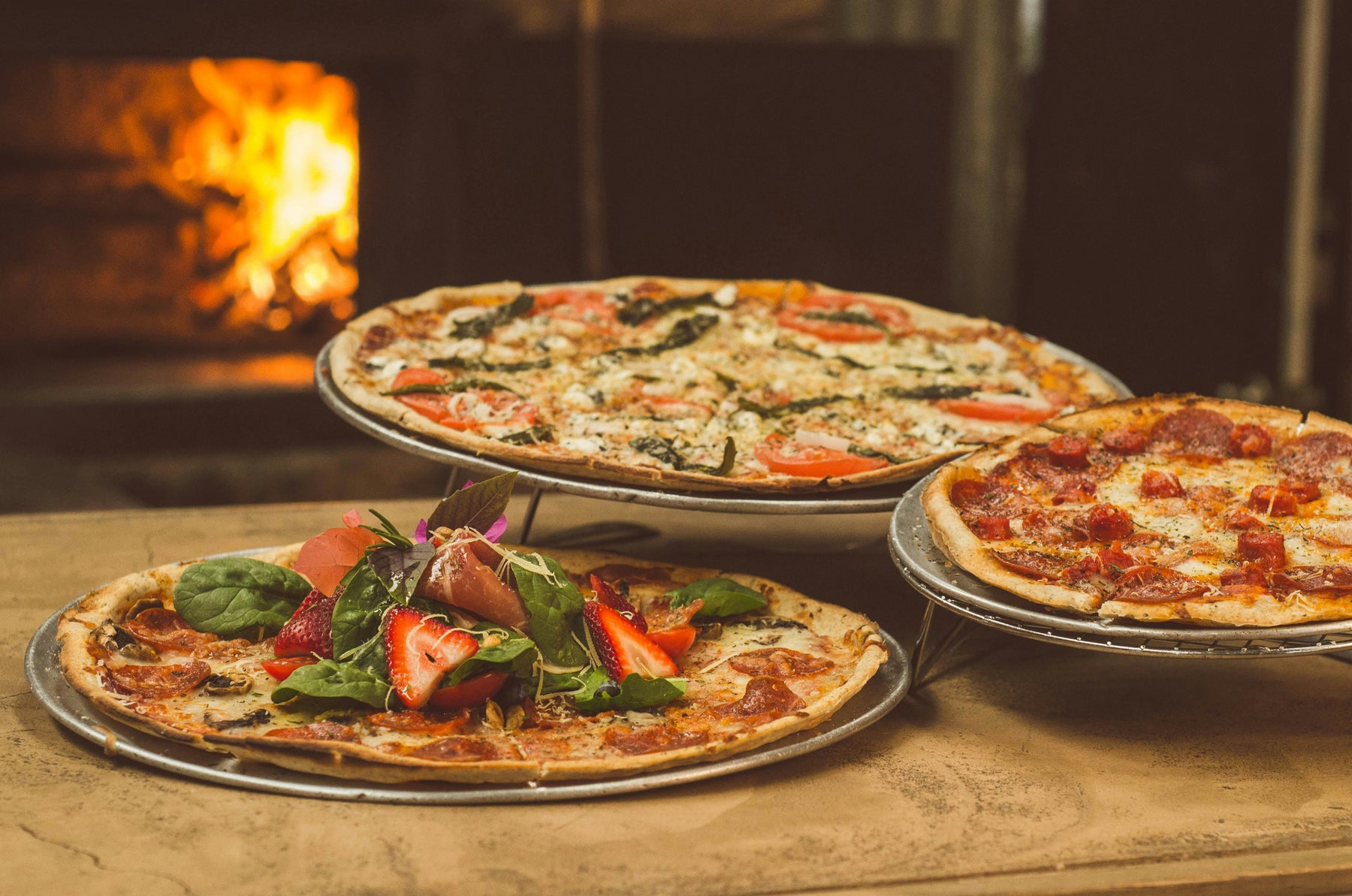 Three gourmet pizzas with unique toppings, including fresh strawberries, greens, and classic pepperoni, served in front of a wood-fired oven.