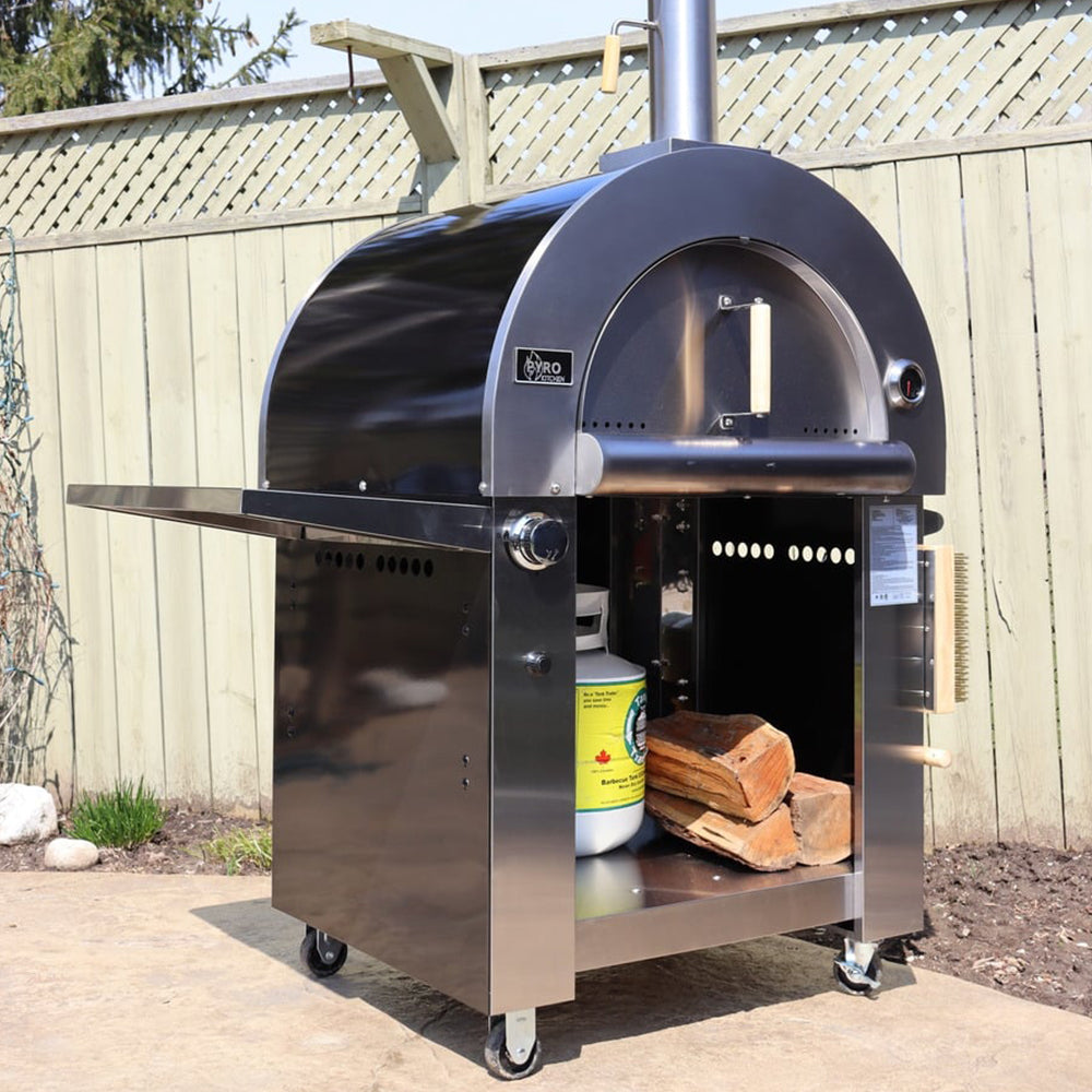 Alternate side view of the Pyro Kitchen Caldo V2 Hybrid Pizza Oven with logs, propane tank, and included accessories like the fold-up peel and log holder.