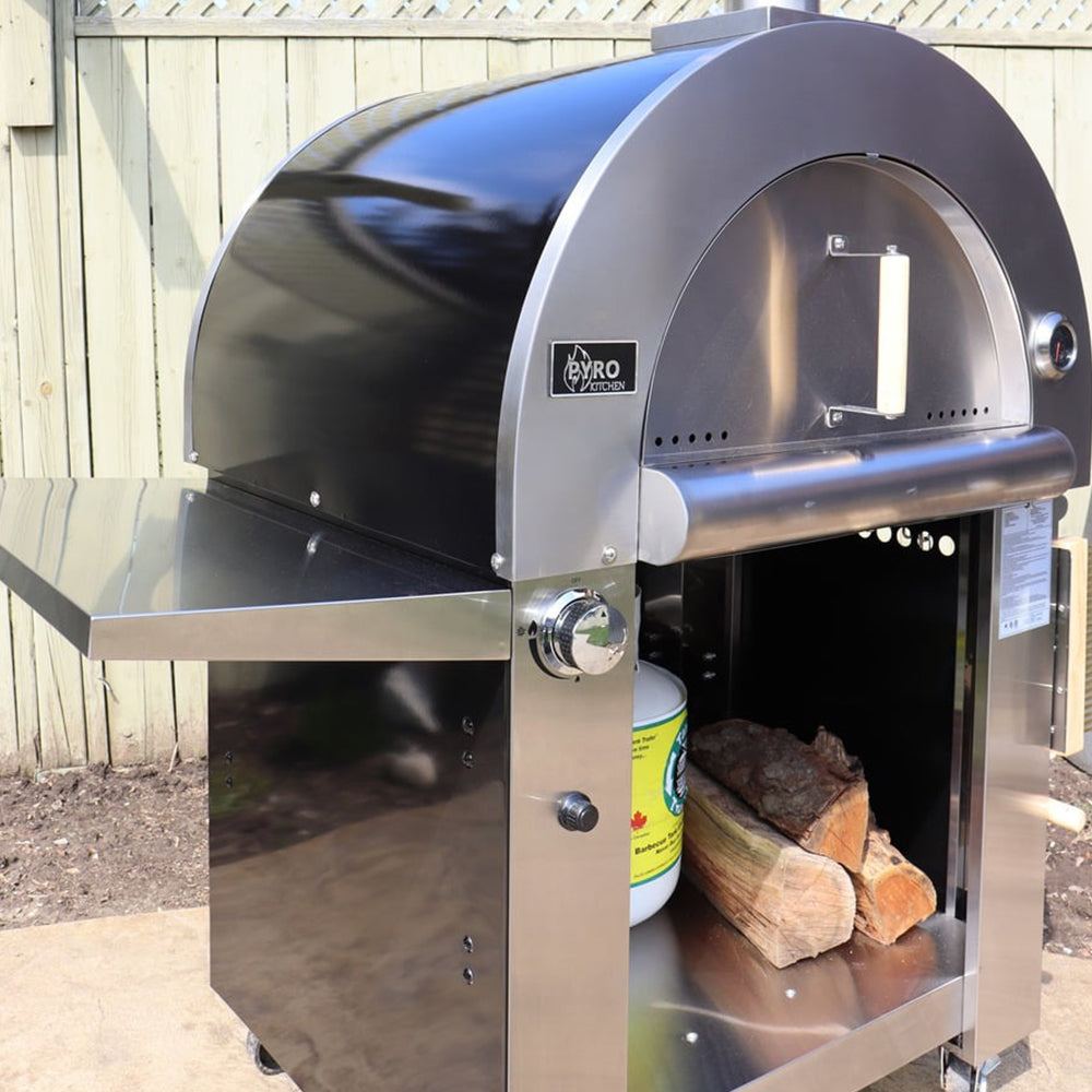 Side view of the Pyro Kitchen Caldo V2 Hybrid Pizza Oven with logs, propane tank, and 14-inch stainless steel side shelf, showcasing hybrid functionality with wood and gas compatibility.
