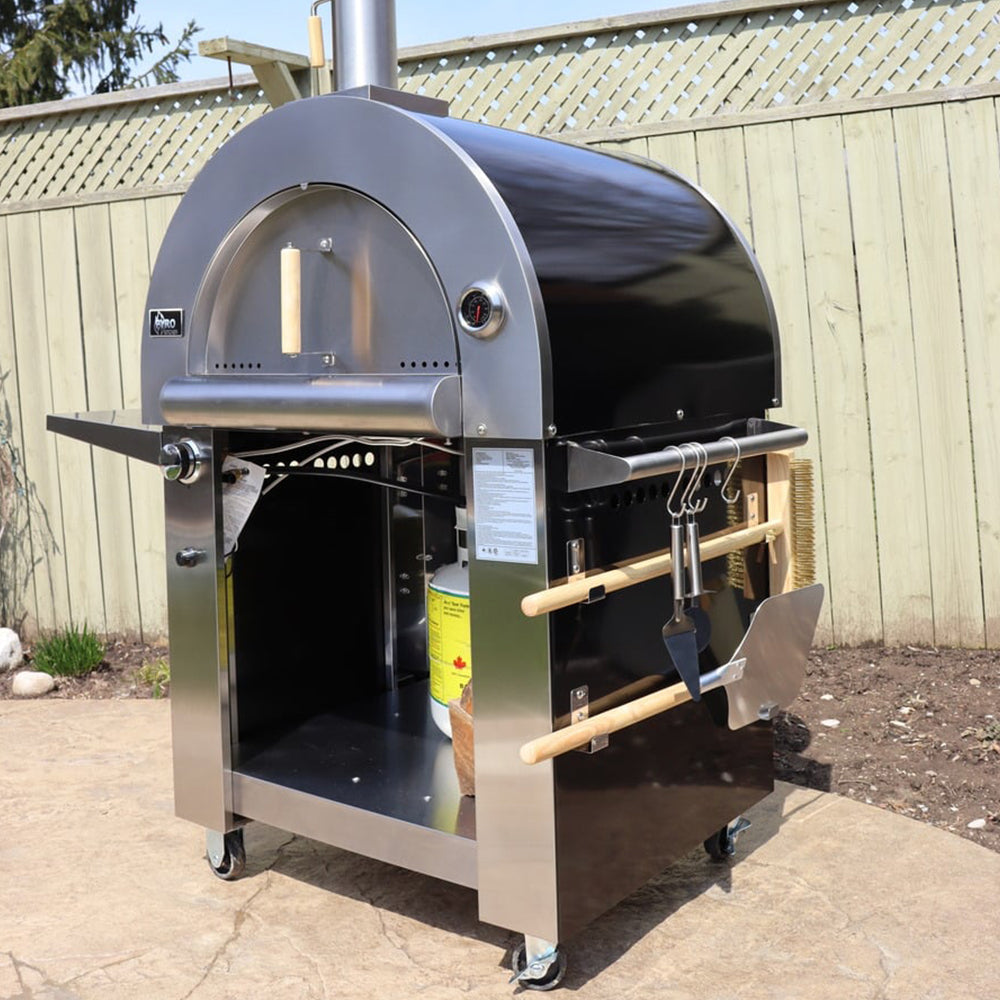 Side view of the Pyro Kitchen Caldo V2 Hybrid Pizza Oven with all accessories, including a fold-up peel, Chicago-style cutter, brush, thick cordierite pizza stones, and log holder.