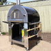 Side view of the Pyro Kitchen Caldo V2 Hybrid Pizza Oven with all accessories, including a fold-up peel, Chicago-style cutter, brush, thick cordierite pizza stones, and log holder.