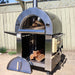 Front view of the Pyro Kitchen Vera V2 Wood Fired Pizza Oven with logs inside the oven and accessories, including pizza peel and brush, on the side shelf.