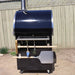 Side view of the Pyro Kitchen Vera V2 Wood Fired Pizza Oven with accessories, including pizza peel, cutter, brush, log holder, and pizza server.